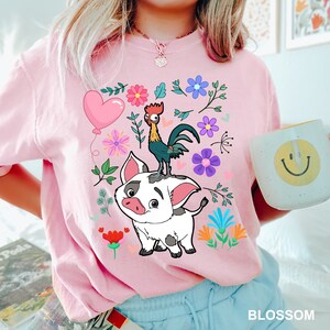 K&ouml;nnte beinhalten: Pinkes T-Shirt mit einem Cartoon-Motiv aus einem Schwein und einem Hahn. Das Schwein ist wei&szlig; mit schwarzen Flecken und der Hahn ist rot, gelb und gr&uuml;n. Das Motiv ist von rosa, lila, blauen und gr&uuml;nen Blumen umgeben.