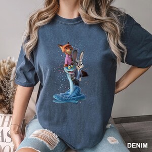 May include: Denim blue t-shirt featuring a colorful graphic of a fox, a rabbit, and a snake. The fox wears a pink shirt, and the rabbit wears a blue suit. The snake is blue. The word "DENIM" is printed on the bottom right.
