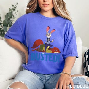 May include: A blue t-shirt featuring a cartoon image of a rabbit and a fox with the word "BUSTED!" in blue. The rabbit is standing on the fox. The shirt is a solid blue color.