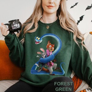May include: Forest green sweatshirt featuring a cartoon graphic of a blue snake wrapped around a fox and a rabbit. The sweatshirt has the words "FOREST GREEN" printed at the bottom.