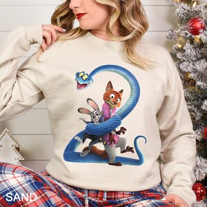 May include: Sand-colored sweatshirt featuring a colorful graphic of Zootopia characters. The design includes Judy Hopps, Nick Wilde, and a blue snake. The word "SAND" is printed at the bottom of the sweatshirt.