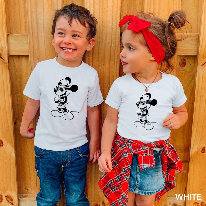 Może przedstawiać: Dwoje dzieci w białych koszulkach z czarno-białym obrazkiem Myszki Miki w okularach przeciwsłonecznych i czapce. Dziewczynka ma na sobie czerwoną kratkowaną sp&oacute;dniczkę i czerwoną opaskę na głowie. Chłopiec ma na sobie niebieskie dżinsy. Tekst "WHITE" jest widoczny w prawym dolnym rogu obrazu.