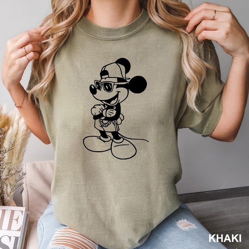 Może przedstawiać: Koszulka w kolorze khaki z czarnym nadrukiem przedstawiającym Myszke Miki w okularach przeciwsłonecznych i czapce z daszkiem. Nadruk przedstawia Myszke Miki z założonymi rękami i złotym łańcuszkiem na szyi.