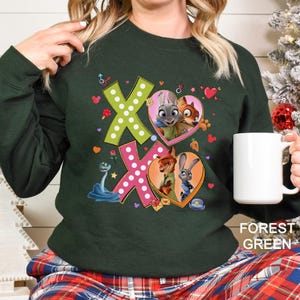 May include: Forest green sweatshirt featuring a colorful graphic of the characters from Zootopia. The design includes the letters "XO" with heart-shaped images of Judy Hopps and Nick Wilde. The sweatshirt is a comfortable, casual style.