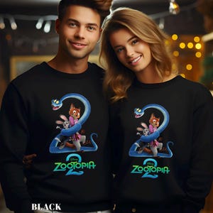 May include: Black sweatshirt featuring a colorful graphic from the movie Zootopia 2. The design includes a blue snake wrapped around cartoon characters. The text "Zootopia 2" is displayed in green and blue. The word "BLACK" is at the bottom.