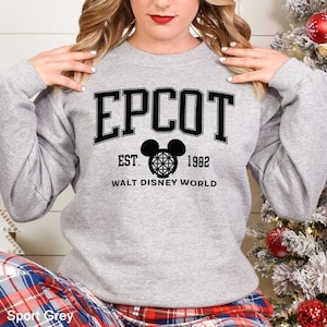 K&ouml;nnte beinhalten: Hellgraues Sweatshirt mit dem Wort "EPCOT" in schwarzen Buchstaben. Darunter ein schwarzes Mickey Mouse Kopf-Design und der Text "EST. 1982 WALT DISNEY WORLD". Das Sweatshirt hat die Farbe Sport Grey.