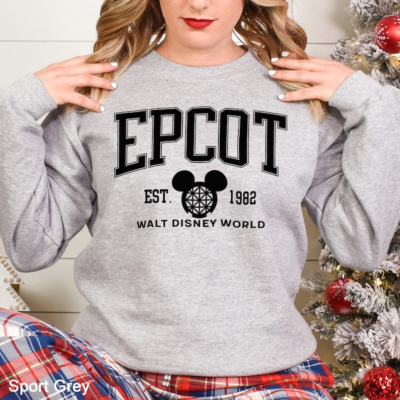 K&ouml;nnte beinhalten: Hellgraues Sweatshirt mit dem Wort "EPCOT" in schwarzen Buchstaben. Darunter ein schwarzes Mickey Mouse Kopf-Design und der Text "EST. 1982 WALT DISNEY WORLD". Das Sweatshirt hat die Farbe Sport Grey.