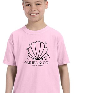 May include: Pink t-shirt with a black graphic of a seashell and the text "Ariel & Co. Since 1989"