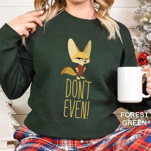 May include: Forest green sweatshirt featuring a cartoon fox illustration and the text "DON'T EVEN!". The fox is wearing a red shirt. A white mug is held in the hand. The sweatshirt is a comfortable, casual style.