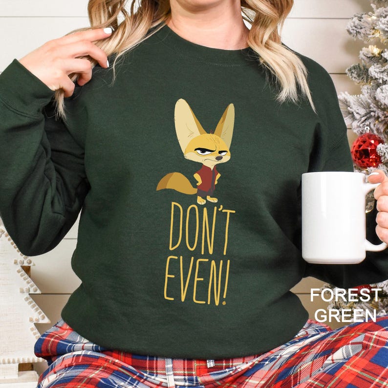 May include: Forest green sweatshirt featuring a cartoon fox illustration and the text "DON'T EVEN!". The fox is wearing a red shirt. A white mug is held in the hand. The sweatshirt is a comfortable, casual style.