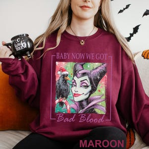 May include: Maroon sweatshirt featuring a graphic of Maleficent with a crow wearing a Santa hat. The text reads "Baby Now We Got Bad Blood" and "Maroon". A black mug with "Witches Brew" is held in the hand.