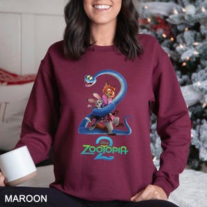 May include: Maroon sweatshirt featuring a graphic of characters from Zootopia 2, including a fox, rabbit, and snake. The Zootopia 2 logo is displayed in green and blue. The sweatshirt is a solid maroon color.