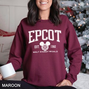 K&ouml;nnte beinhalten: Burgunderfarbenes Sweatshirt mit dem Wort "EPCOT" in wei&szlig;en Blockbuchstaben. Darunter steht der Text "EST. 1982" und "WALT DISNEY WORLD". Eine Mickey Mouse-Kopf-Grafik ist ebenfalls vorhanden.