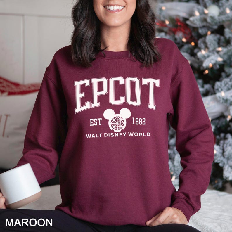 K&ouml;nnte beinhalten: Burgunderfarbenes Sweatshirt mit dem Wort "EPCOT" in wei&szlig;en Blockbuchstaben. Darunter steht der Text "EST. 1982" und "WALT DISNEY WORLD". Eine Mickey Mouse-Kopf-Grafik ist ebenfalls vorhanden.