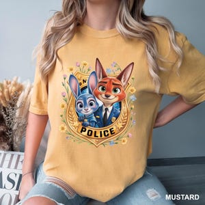 K&ouml;nnte beinhalten: Senffarbenes T-Shirt mit einem Cartoon-Bild eines Hasen und eines Fuchses in Polizeiuniformen in einem goldenen Abzeichen. Das Abzeichen ist von Blumen und dem Wort "POLICE" umgeben. Das Shirt ist ein l&auml;ssiger Stil.