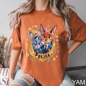 K&ouml;nnte beinhalten: Gebrannt orangefarbenes T-Shirt mit einem Cartoon-Bild eines blauen Kaninchens und eines roten Fuchses in Polizeiuniformen, umgeben von Blumen und einem goldenen Abzeichen mit der Aufschrift "POLICE". Das T-Shirt hat kurze &Auml;rmel.