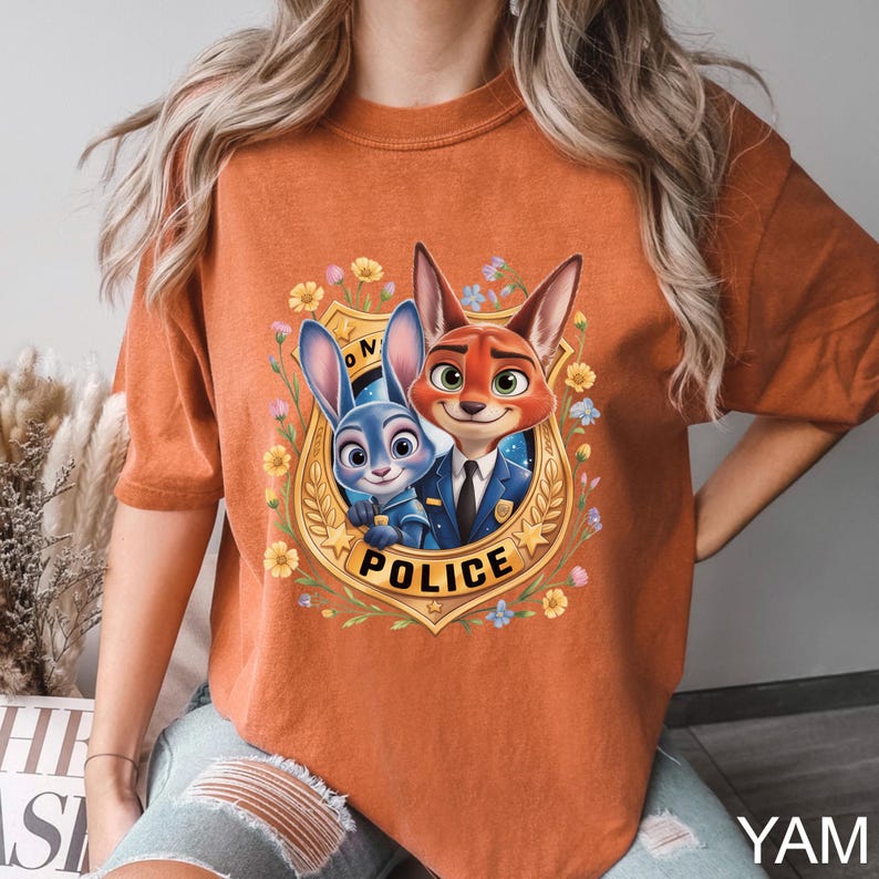 K&ouml;nnte beinhalten: Gebrannt orangefarbenes T-Shirt mit einem Cartoon-Bild eines blauen Kaninchens und eines roten Fuchses in Polizeiuniformen, umgeben von Blumen und einem goldenen Abzeichen mit der Aufschrift "POLICE". Das T-Shirt hat kurze &Auml;rmel.