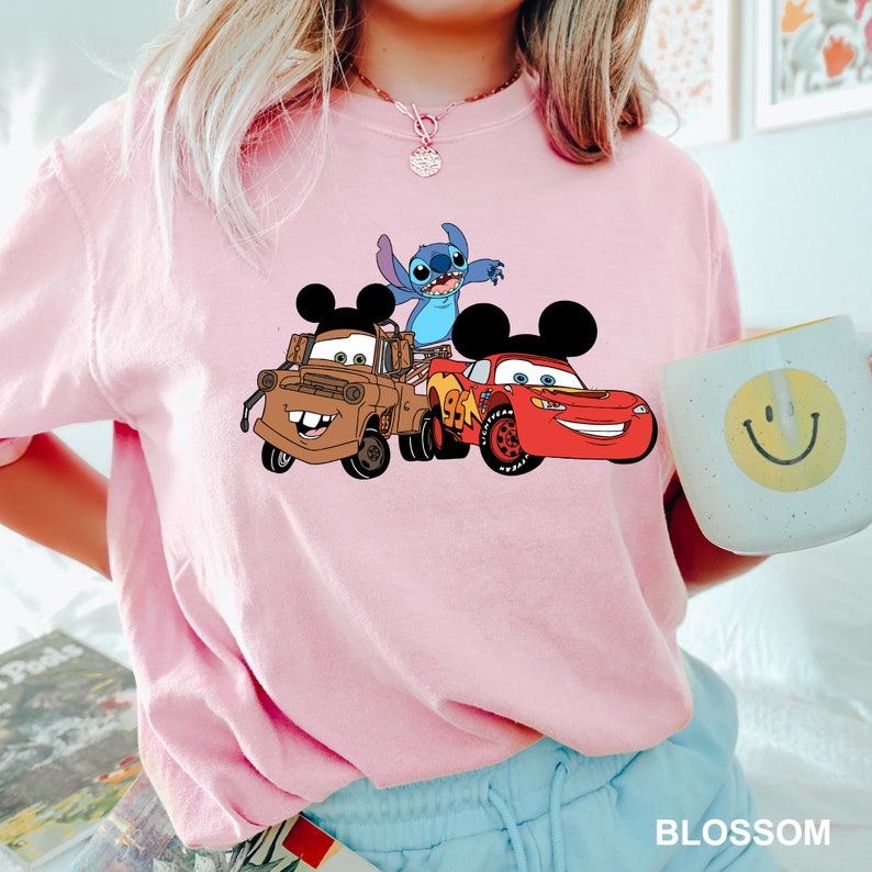 May include: Pink t-shirt with a graphic of Stitch, Mickey Mouse, and Lightning McQueen from Disney's Cars. The characters are in a playful pose, with Stitch on top of Mickey Mouse's head and Lightning McQueen driving in front.