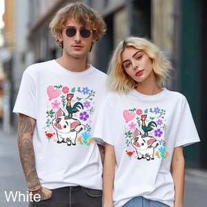 K&ouml;nnte beinhalten: Wei&szlig;es T-Shirt mit einem bunten Blumendesign, das einen Hahn zeigt, der auf einem Schwein reitet. Das Design enth&auml;lt einen rosafarbenen Herzluftballon und gr&uuml;ne Bl&auml;tter.