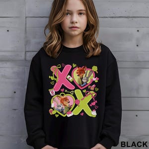 May include: Black sweatshirt featuring a colorful graphic of characters from the movie Zootopia. The design includes the letters "X" and "O" with heart-shaped frames, ribbons, and cupcakes. The word "BLACK" is printed at the bottom right.
