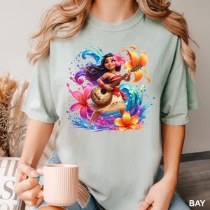 Puede incluir: Camiseta verde claro con una colorida ilustraci&oacute;n de una mujer polinesia tocando el ukelele. La obra de arte incluye flores vibrantes, salpicaduras de agua y la palabra "BAY". La mujer lleva un top rojo y una falda de hierba.