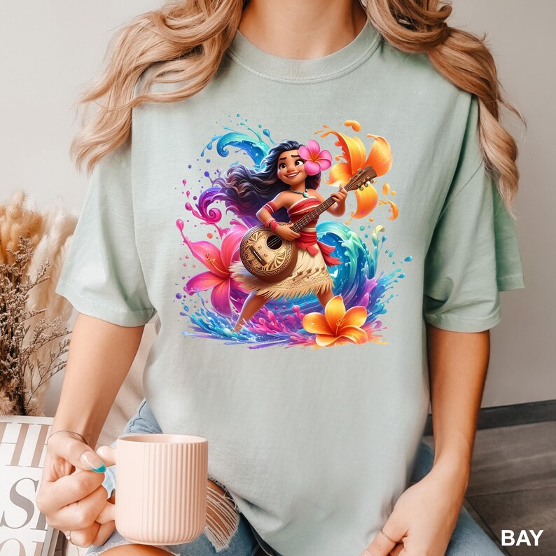 Puede incluir: Camiseta verde claro con una colorida ilustraci&oacute;n de una mujer polinesia tocando el ukelele. La obra de arte incluye flores vibrantes, salpicaduras de agua y la palabra "BAY". La mujer lleva un top rojo y una falda de hierba.