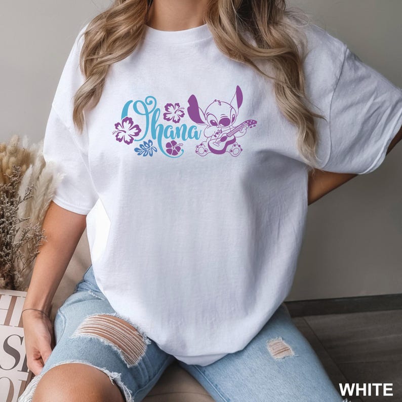 Puede incluir: Camiseta blanca con la palabra "Ohana" en escritura turquesa y morada, detalles florales y un personaje de dibujos animados tocando la guitarra. La camiseta es de material suave.