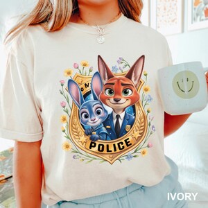 K&ouml;nnte beinhalten: Cremefarbenes T-Shirt mit einem Cartoon-Bild eines blauen Kaninchens und eines roten Fuchses in Polizeiuniformen, eingebettet in ein goldenes Abzeichen mit der Aufschrift "POLIZEI". Das Design ist von bunten Blumen umgeben. Ein wei&szlig;er Becher mit einem Smiley wird in der N&auml;he gehalten.