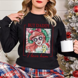 May include: Black sweatshirt featuring a graphic of a cartoon character with red hair and sunglasses, and a yellow character wearing a Santa hat. The text "BUT DADDY I love him" is printed in red. The word "BLACK" is printed at the bottom.