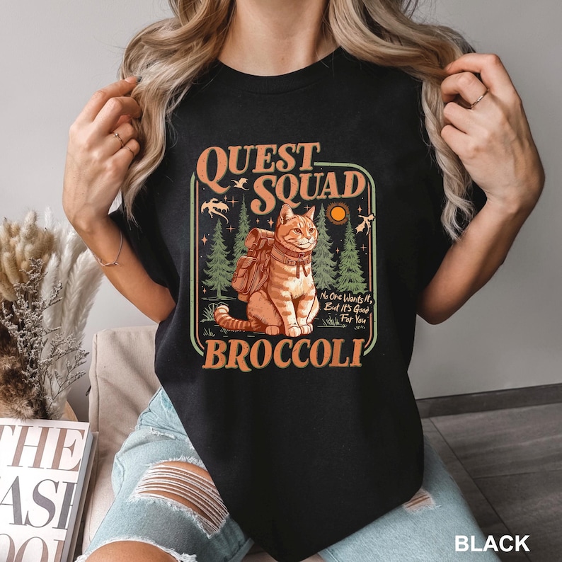 May include: Black t-shirt featuring a graphic of a cat with a backpack, the words "Quest Squad" and "Broccoli". The design includes a forest scene and the text "No One Wants It, But It's Good For You".