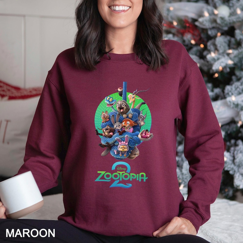 May include: Maroon sweatshirt featuring a colorful graphic of characters from the animated movie "Zootopia 2". The design includes a green circle with various animal characters, the movie title, and the word "MAROON" at the bottom.