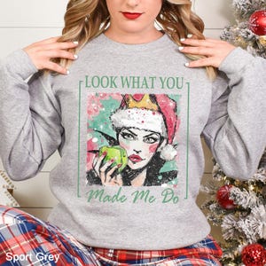 May include: Sport Grey sweatshirt featuring a graphic of a woman wearing a Santa hat and holding a green apple. The text "LOOK WHAT YOU MADE ME DO" is printed above the graphic, and "Made Me Do" is printed below. The sweatshirt is a light gray color.