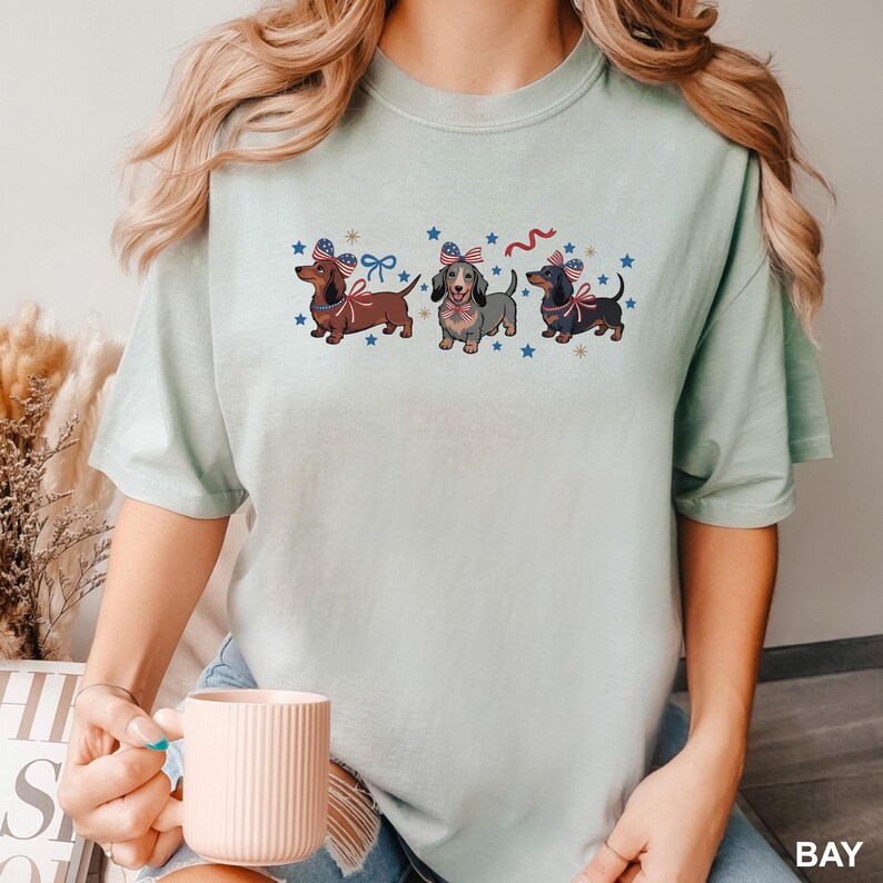 May include: A light green t-shirt featuring three dachshunds with patriotic bows and surrounded by blue stars. The dachshunds are in shades of brown, gray, and black. The shirt has a relaxed fit and short sleeves.
