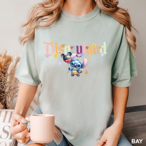 Puede incluir: Una camiseta azul claro con un gr&aacute;fico colorido de "Disneyland" que presenta a Stitch sosteniendo un globo de Mickey Mouse. El gr&aacute;fico est&aacute; rodeado de peque&ntilde;as flores.