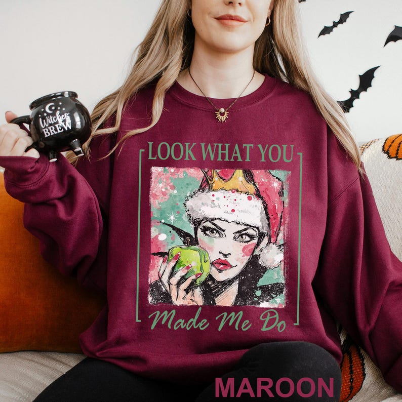 May include: Maroon sweatshirt with a graphic of a woman wearing a Santa hat and holding a green apple. The text "LOOK WHAT YOU MADE ME DO" and "MAROON" are printed on the sweatshirt. A black mug with the text "Witches Brew" is held in the left hand.