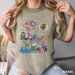 May include: Khaki-colored t-shirt featuring a colorful graphic of characters from the animated movie Zootopia. The design includes various animal characters in frames and the word "Zootopia". The shirt is a casual style.
