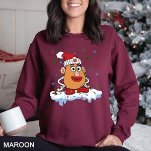 Puede incluir: Sudadera granate con un personaje de dibujos animados de Mr. Potato Head con un gorro de Pap&aacute; Noel y luces navide&ntilde;as. El dise&ntilde;o incluye boca, ojos y orejas rojas. La palabra "MAROON" est&aacute; impresa en la parte inferior de la imagen.