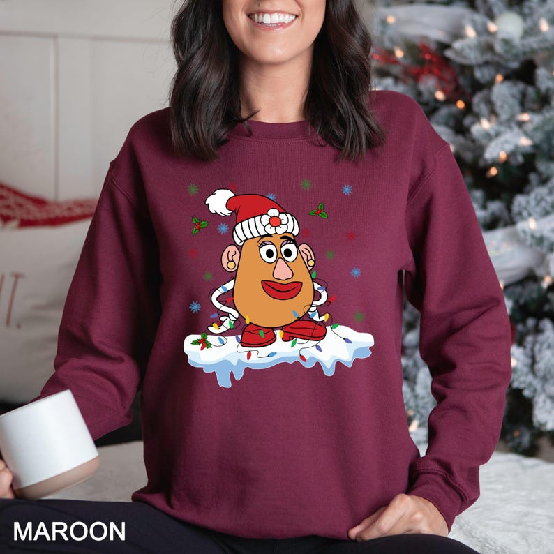 Puede incluir: Sudadera granate con un personaje de dibujos animados de Mr. Potato Head con un gorro de Pap&aacute; Noel y luces navide&ntilde;as. El dise&ntilde;o incluye boca, ojos y orejas rojas. La palabra "MAROON" est&aacute; impresa en la parte inferior de la imagen.