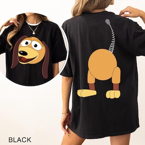 May include: Black t-shirt with a cartoon dog printed on the front and the back. The dog on the front is facing forward with its mouth open. The dog on the back is facing away from the viewer with its tail curled up. The text "BLACK" is printed below the image.