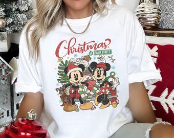 May include: White t-shirt featuring Mickey and Minnie Mouse in Christmas attire. The graphic includes the word "Christmas" in red script, and a street sign that reads "Main Street". The characters are surrounded by Christmas decorations.