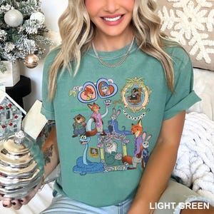 May include: Light green t-shirt featuring cartoon characters from Zootopia. The design includes framed images of the characters, the word "Zootopia", and the word "Wootopia". The shirt is a solid light green color.