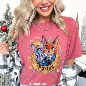 K&ouml;nnte beinhalten: Karmesinrotes T-Shirt mit einem Cartoon-Bild eines Fuchses und eines Hasen in Polizeiuniformen. Das Bild ist von einem goldenen Abzeichen mit dem Wort "POLICE" und floralen Akzenten umgeben. Das Shirt hat einen Rundhalsausschnitt und kurze &Auml;rmel.