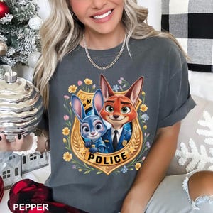 K&ouml;nnte beinhalten: Dunkelgraues T-Shirt mit einem Cartoon-Bild eines Hasen und eines Fuchses in Polizeiuniformen, umgeben von Blumen und dem Wort "POLICE". Das T-Shirt hat einen Rundhalsausschnitt und kurze &Auml;rmel.