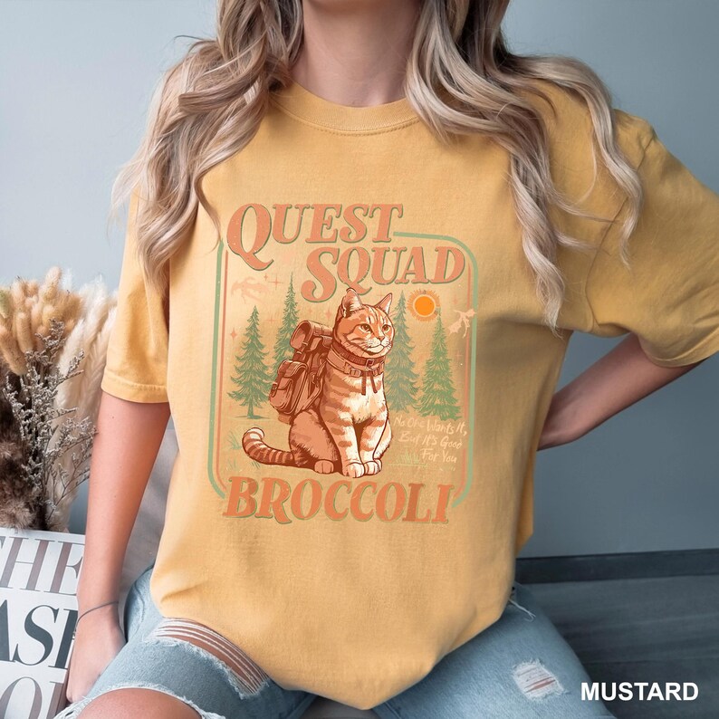 May include: Mustard-colored t-shirt featuring a graphic of a cat with a backpack, the words "Quest Squad Broccoli", and a nature scene. The design includes trees, a sun, and the phrase "No One Wants It, But It's Good For You".