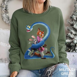 May include: Military green sweatshirt featuring a cartoon graphic of characters from Zootopia, including Judy Hopps, Nick Wilde, and a blue snake. The design is centered on the front of the long-sleeved top.