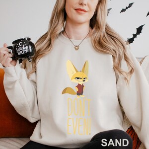 May include: Sand-coloured sweatshirt with a cartoon fox in a red vest and an angry expression. The text "DON'T EVEN!" is printed in yellow below the fox. The word "SAND" is printed at the bottom of the sweatshirt.