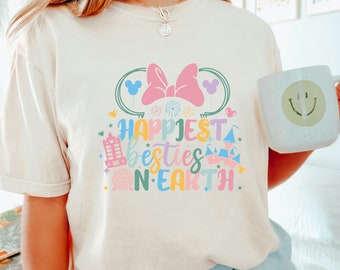 Op de afbeelding: Een wit T-shirt met een roze strik en de tekst "Happiest besties on Earth" in kleurrijke letters. Het ontwerp bevat een Mickey Mouse-hoofd, een kasteel en andere Disney-thema-iconen.