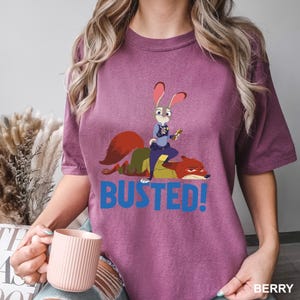 May include: Berry-colored t-shirt featuring a cartoon image of a rabbit police officer and a fox with the word "BUSTED!" in blue. The rabbit is wearing a police uniform and holding a small object. The shirt is a comfortable fit.