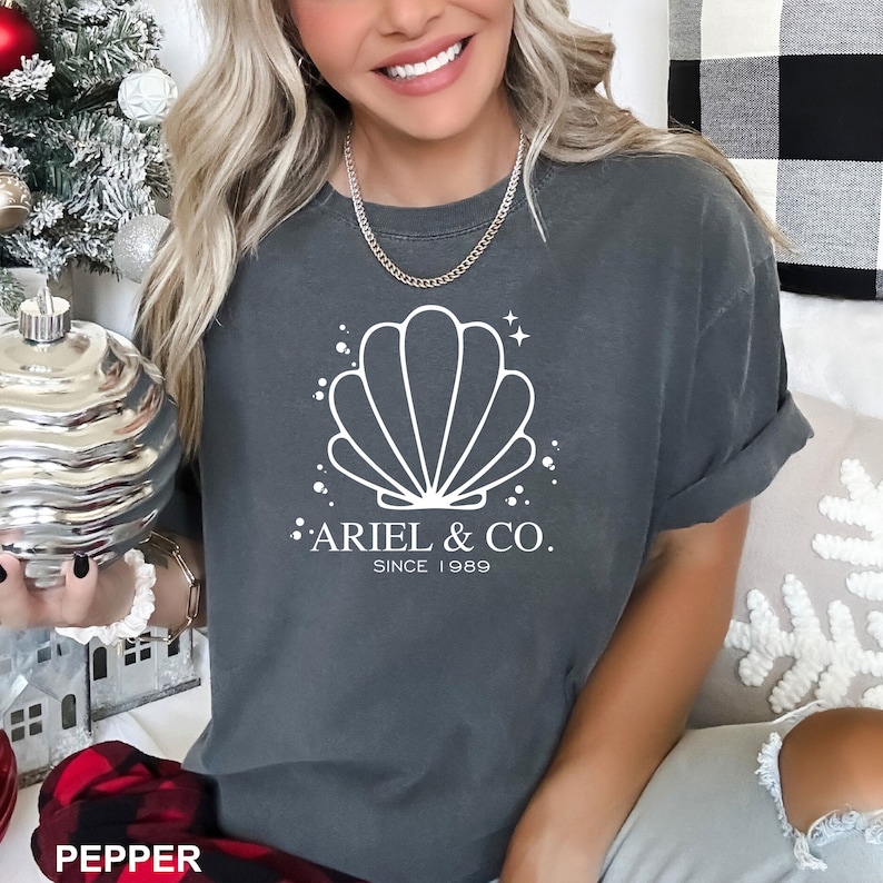 May include: A dark gray t-shirt with a white seashell graphic and the text "Ariel & Co. Since 1989".