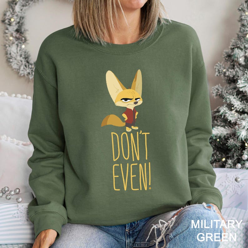 May include: Military green sweatshirt with a cartoon fox illustration. The fox has an angry expression and wears a red vest. The text "DON'T EVEN!" is in yellow below the fox. The sweatshirt is a solid colour.